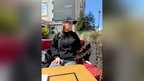 Young woman in a black sweatshirt and cap sitting outdoors on a sunny day near a garden planter.