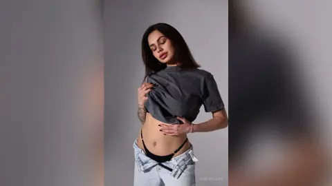 Young woman in a grey cropped t-shirt and light denim jeans posing confidently against a grey studio background.