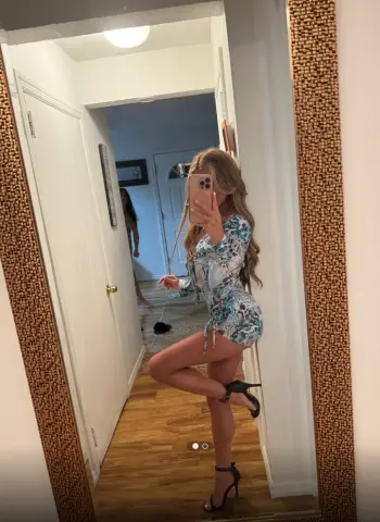 Blonde woman in a blue floral mini dress posing for a mirror selfie in a bright hallway.