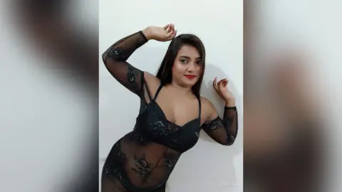 Young woman in a black lace bodysuit posing confidently against a white wall.