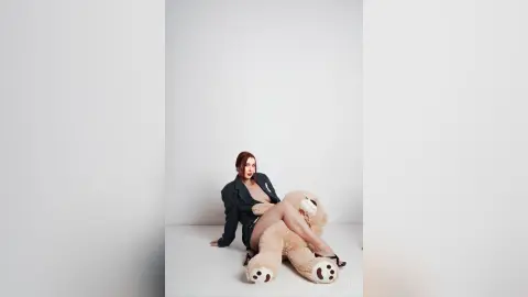 AryaSin sits on a white floor wearing a dark grey blazer, posing with a large tan teddy bear against a plain white background.