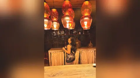 Elegant woman in a black lace dress sitting in a moody restaurant with warm glowing pendant lights.
