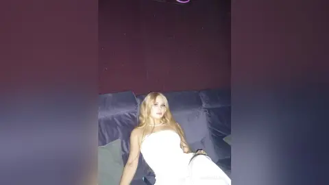 KrystalLaaz is a blonde woman in a white dress lounging on a dark grey couch.