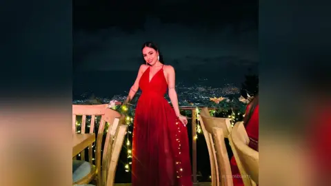 Smiling woman in a long red evening gown posing on a balcony overlooking city lights at night.
