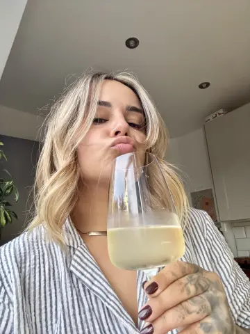 Blonde woman in a striped shirt sipping white wine in a bright kitchen with a playful mood.