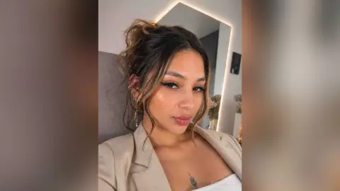 Young woman with messy bun and gold earrings wearing a beige blazer in a modern lit room.