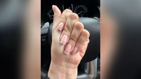 Close-up of elegant French tip manicure on a woman's hand resting against a black car steering wheel.
