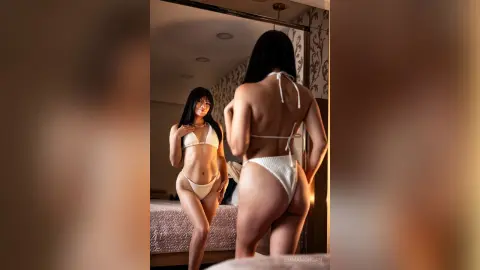 Young woman in a white bikini posing for a mirror selfie in a cozy, sunlit bedroom.