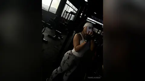 PamelaCreed poses in a dark gym wearing a white tank top and patterned grey leggings while taking a mirror selfie.