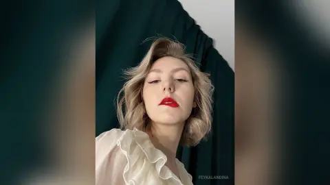 Elegant woman in a white ruffled blouse with bold red lipstick posing against a dark green curtain backdrop.