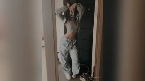 TarenLeonetti poses in a mirror wearing a light grey crop top and matching sweatpants, showing her flat stomach and midriff.