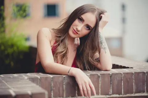 Young woman with long hair in a red tank top leaning on a brick wall in a soft outdoor setting.