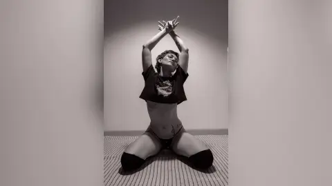 IveteDurand poses with her arms raised above her head, wearing a black t-shirt and black thong while kneeling on a textured floor.