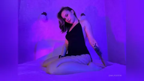 EmilyGiffin wearing a black tank top and silver body chain, sitting on a bed under purple light.