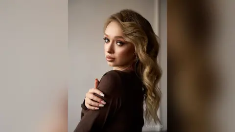 Elegant woman with wavy blonde hair in a dark brown blouse posing with a moody, sophisticated expression.