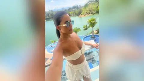 Smiling woman in white bikini and sunglasses relaxing on a sunny tropical resort balcony overlooking a blue lake.