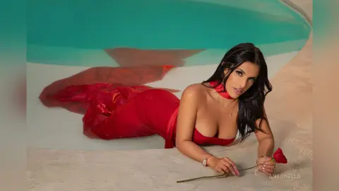Glamorous woman in a flowing red evening gown lying by a turquoise pool holding a single red rose.