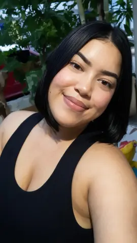 AlejaGoldk smiles while wearing a black tank top against a background of green leaves.