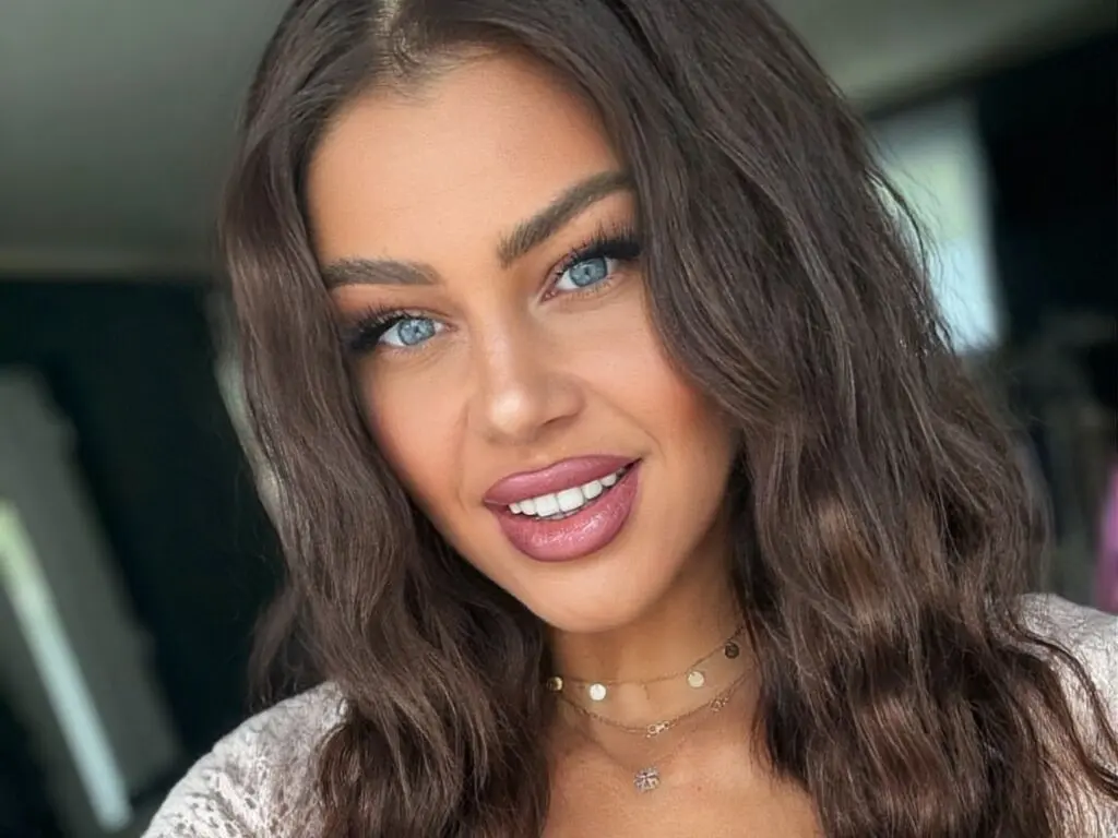 Smiling young woman with wavy brown hair and blue eyes wearing a cozy white knit sweater in soft natural light.