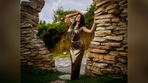 Elegant woman in a shimmering gold satin outfit posing between rustic stone walls in a lush garden setting.