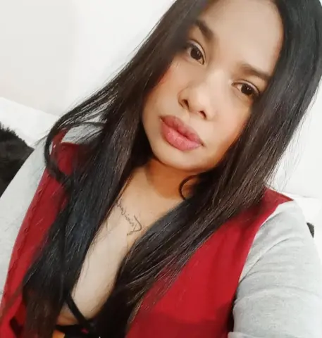 Young woman with long dark hair wearing a red vest and grey shirt in a bright indoor setting.