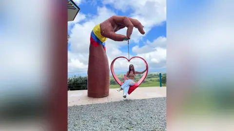 Woman in light blue jeans sitting on a heart-shaped swing held by a giant hand sculpture under a blue sky.