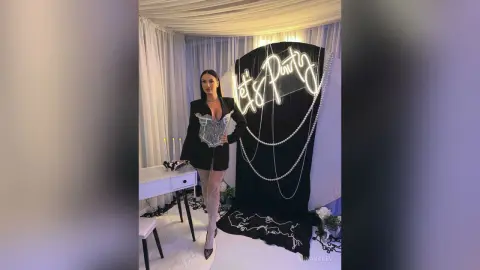 AnnaKarev poses in a black blazer and sheer lace tights standing next to a Let's Party sign.