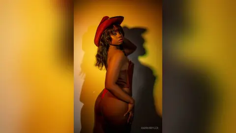 Glamorous woman in red cowboy hat and lingerie posing against a warm yellow background with dramatic shadows.