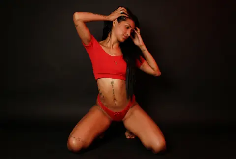 Tattooed woman in a red crop top and bikini bottoms posing against a dark studio background.