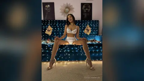 Young woman in white lingerie and gold heels sitting on a blue velvet sofa in a moody, dimly lit room.