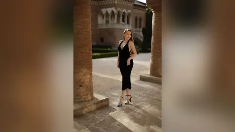 RubySoul stands outdoors in a black sleeveless dress and high heels, posing between two brick pillars on a stone path.