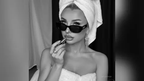 Glamorous woman in a white spa towel and sunglasses applying lipstick in a moody black and white portrait.