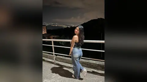 Young woman in a black tank top and blue jeans standing on a balcony overlooking a city at night.