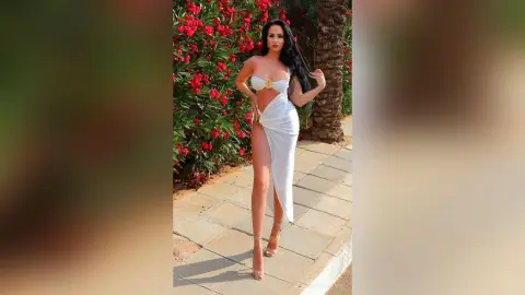 Woman in a white cut-out maxi dress posing in a sunny garden with red flowers.