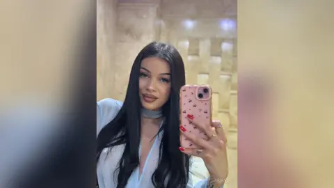 AriaVex takes a mirror selfie wearing a white button-down shirt and a light blue choker, showing off her long black hair.