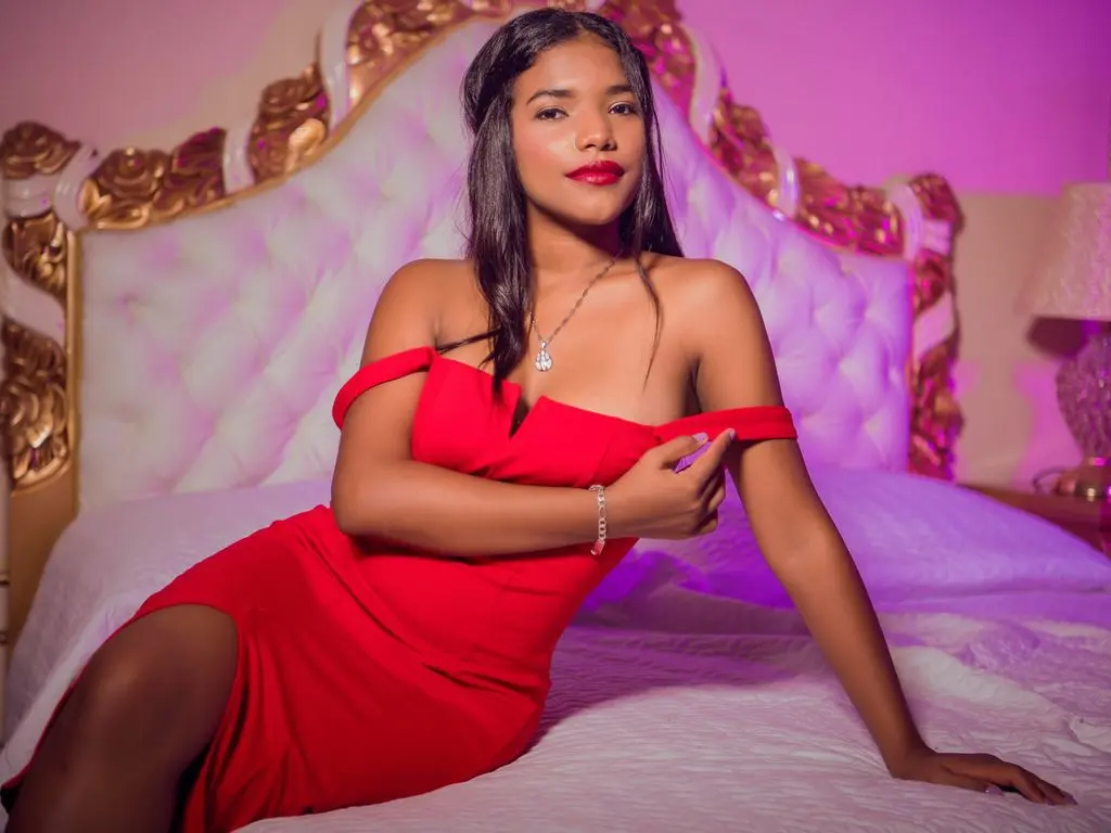 Elegant woman in a red cocktail dress sitting on a bed in a romantic, dimly lit bedroom setting.