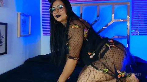 Woman in sheer black floral bodysuit posing on a bed with moody blue neon lighting.