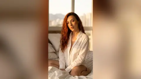 Redheaded woman in a white button-down shirt sitting on a bed in warm, sunlit bedroom lighting.