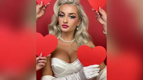 Glamorous blonde woman in a white satin dress and pearls holding red paper hearts against a red background.