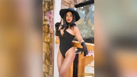 Stylish woman in a black bodysuit and wide-brimmed hat posing confidently on a balcony at sunset.