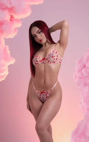 Red-haired woman in red heart print bikini posing against dreamy pink cotton candy clouds.