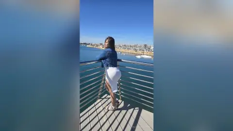 Woman in a denim jacket and white skirt posing on a sunny pier overlooking a coastal city skyline.