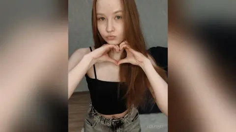 Redheaded woman in a black crop top making a heart gesture with her hands in a cozy indoor setting.