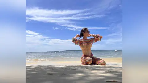 LucyMartin kneels on a sandy beach wearing a colorful crocheted bikini top and matching skirt under a blue sky.
