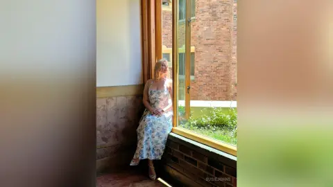 Blonde woman in a floral maxi dress leaning by a sunlit window in a rustic interior setting.