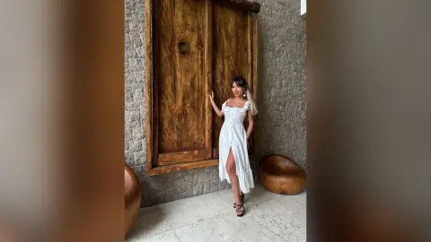 Katia poses in a white sleeveless dress and black heels against a rustic wooden door and stone wall.