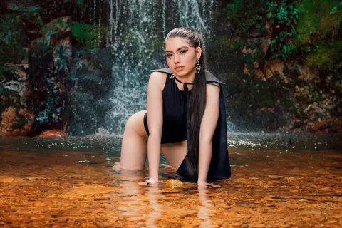 Elegant woman in a black sleeveless outfit posing in a shallow forest waterfall with a serene mood.