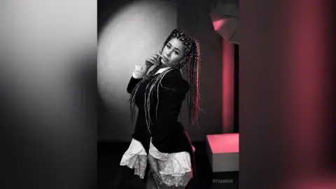 Stylish woman with long braids in a black blazer and white skirt posing in a moody studio setting.