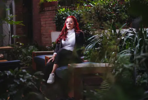 Red-haired woman in a white turtleneck and black blazer sitting in a lush, moody garden setting.