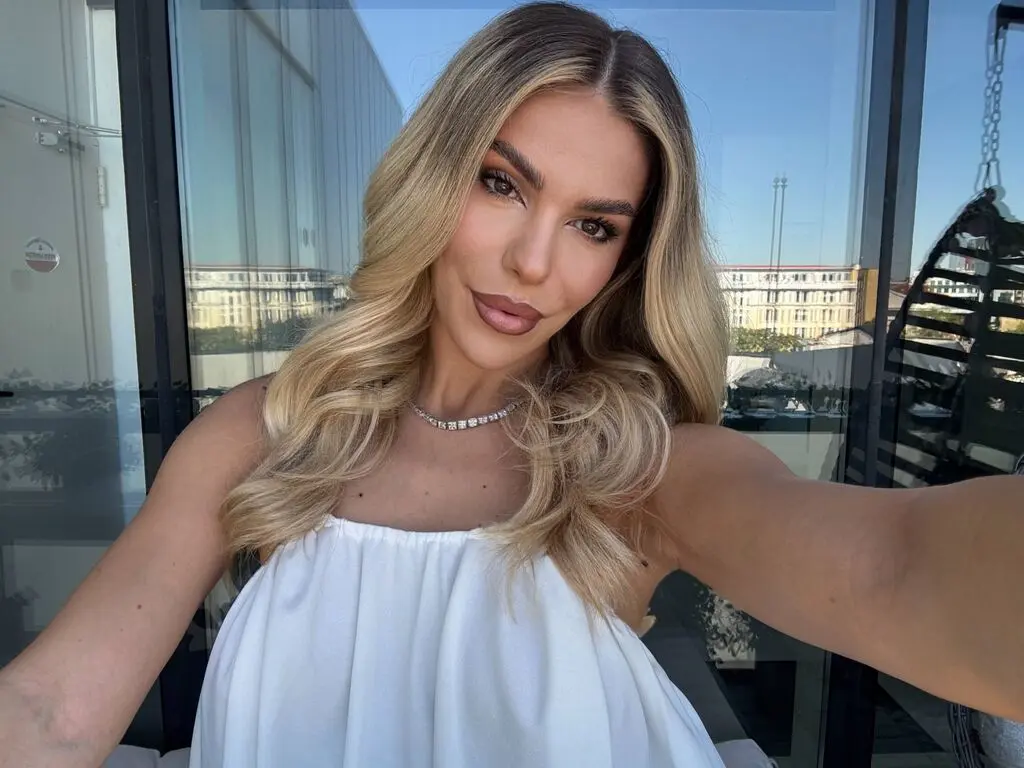 Smiling blonde woman in a white top wearing a silver necklace on a sunny balcony.